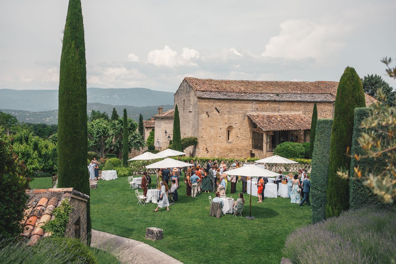 Outdoor wedding reception on lush green lawn beside rustic stone villa in Tuscany with cypress trees and mountain views.