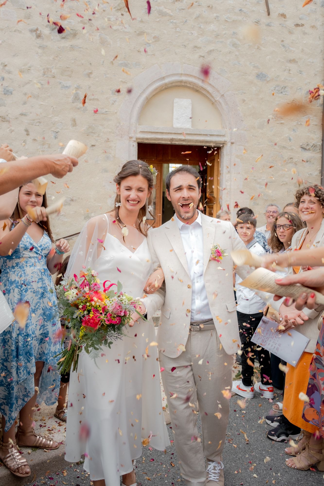 Newlywed couple exits church amid colorful confetti shower, bride holding bouquet, groom in beige suit, guests cheering.