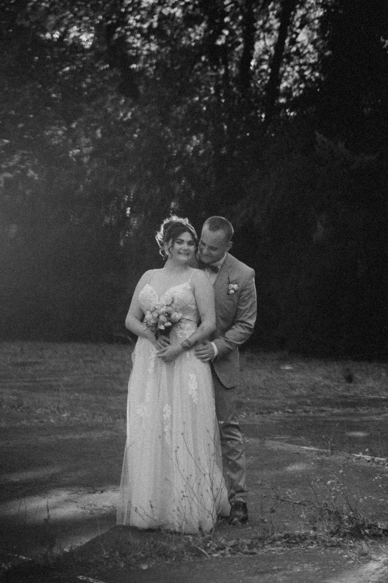 Black and white wedding photo of bride holding bouquet and groom embracing her outdoors near trees.