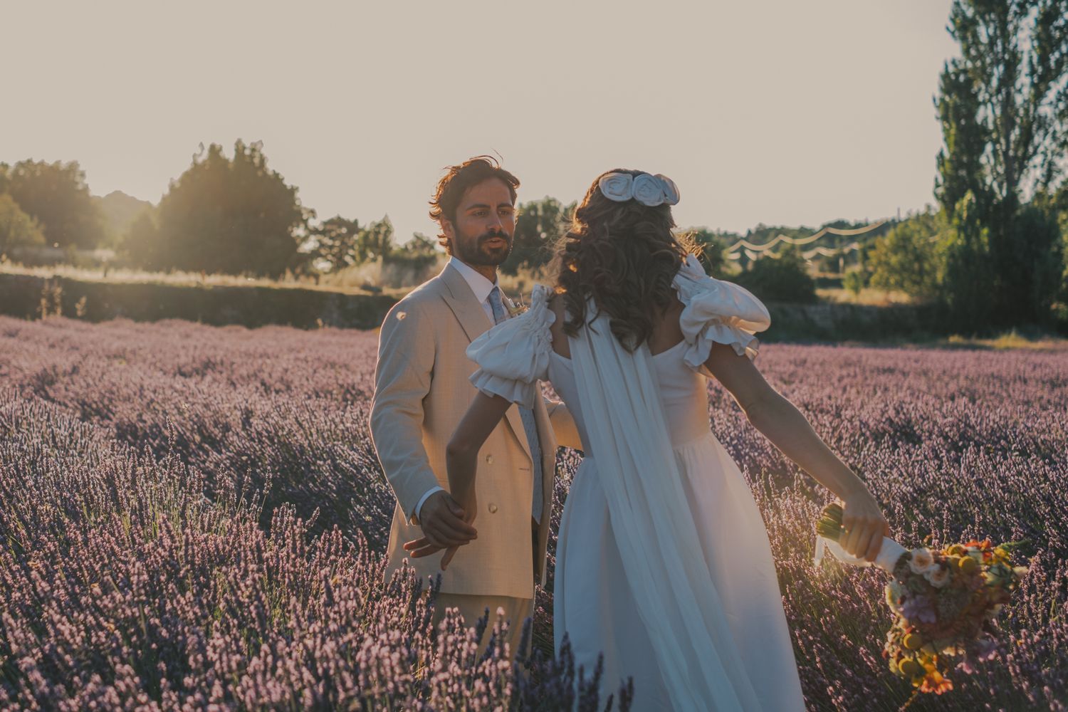 Couple in wedding attire standing in a sunlit lavender field, bride holding wildflowers, golden hour glow.