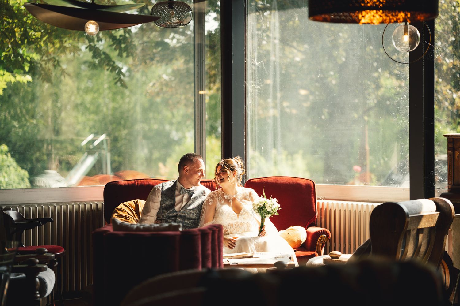 Bride and groom sitting together on a red sofa in an elegant restaurant with large windows and lush greenery outside.