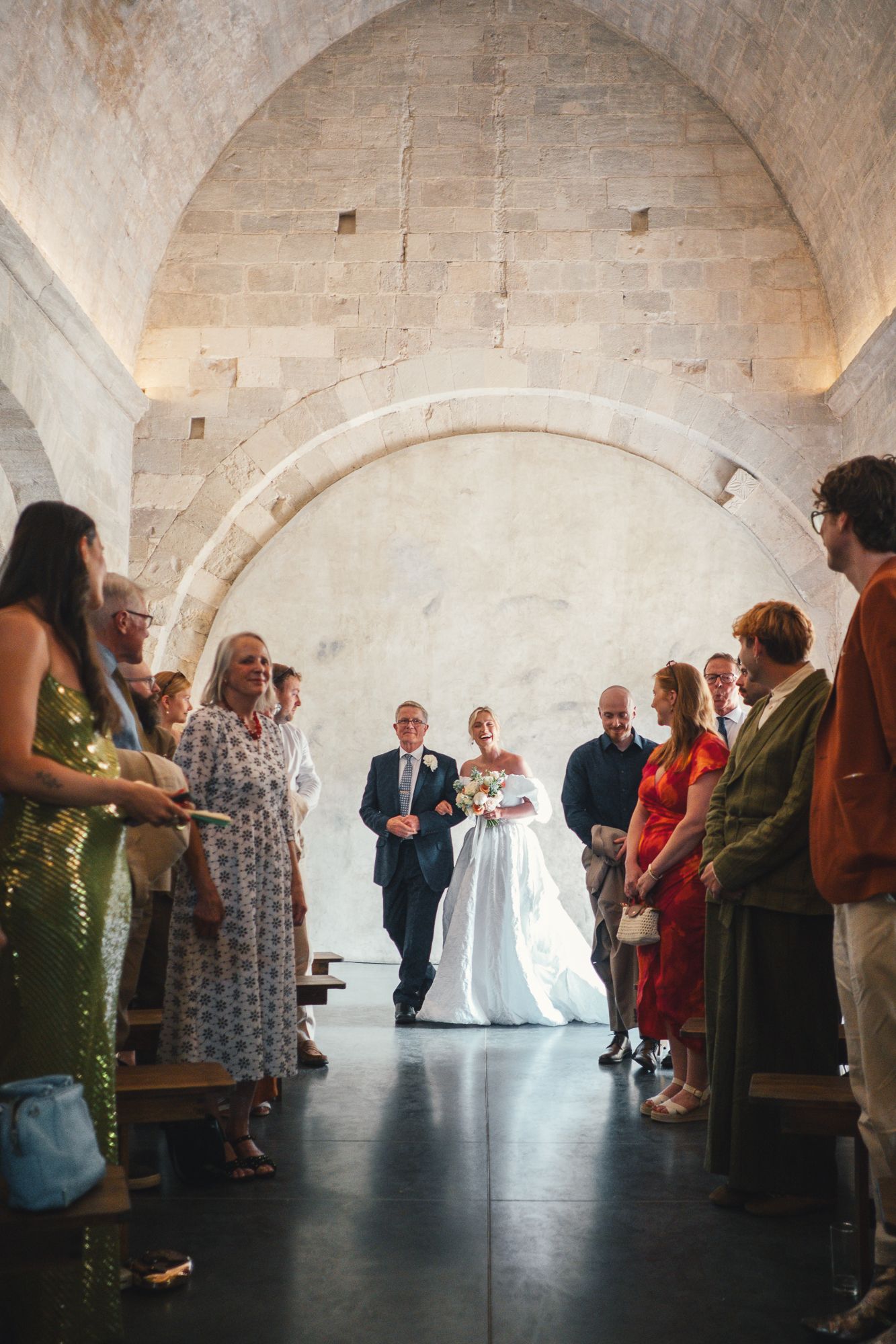 Bride walks down the aisle with escort in a stunning stone arch venue, guests lining the path.