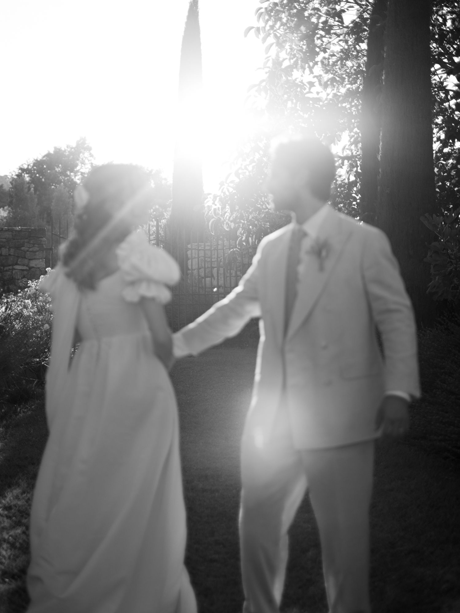 Black and white photo of bride and groom holding hands walking toward sunlit garden gate.