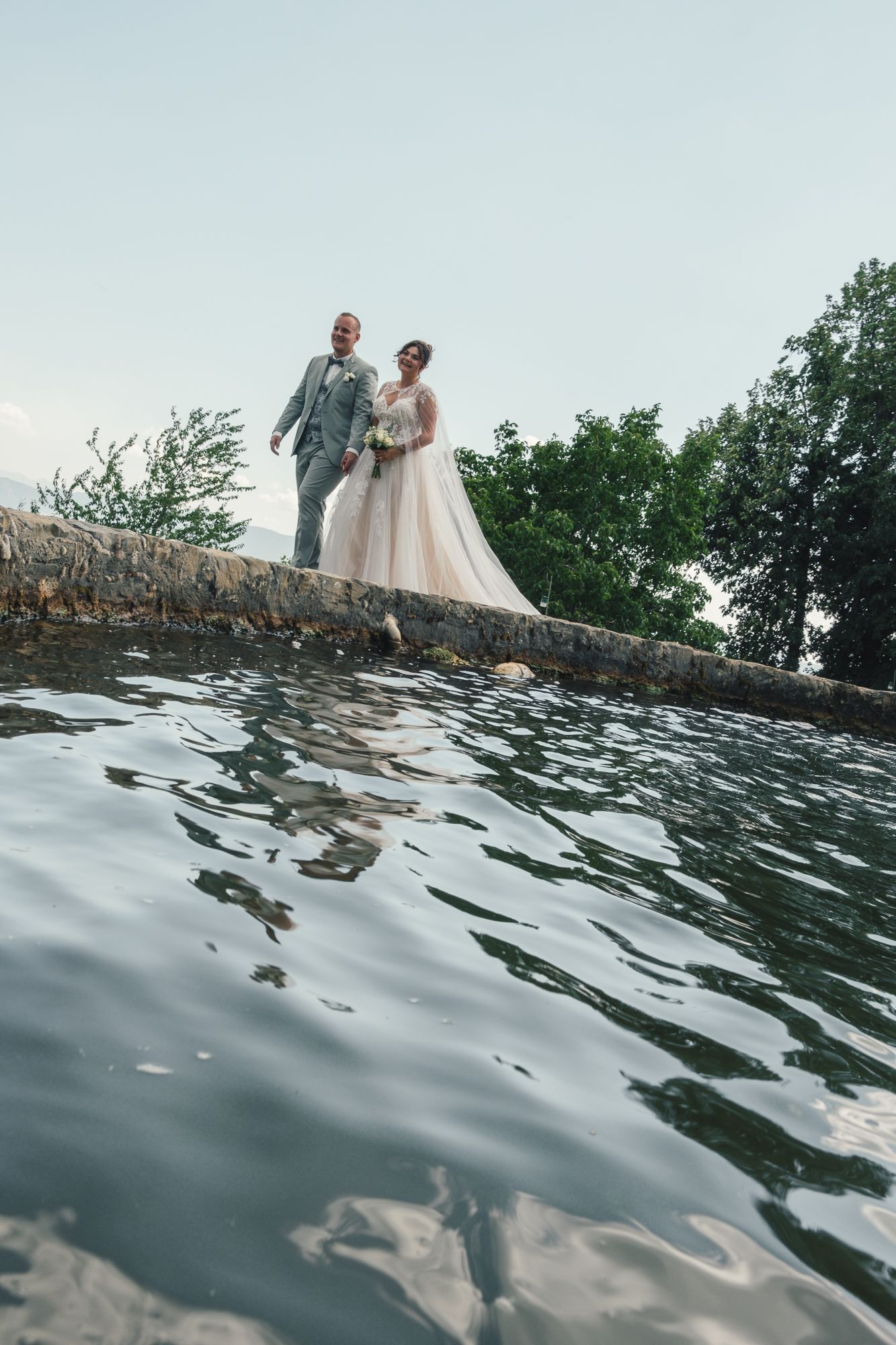 Bride and groom standing on a stone wall by a lake, surrounded by lush green trees on their wedding day.