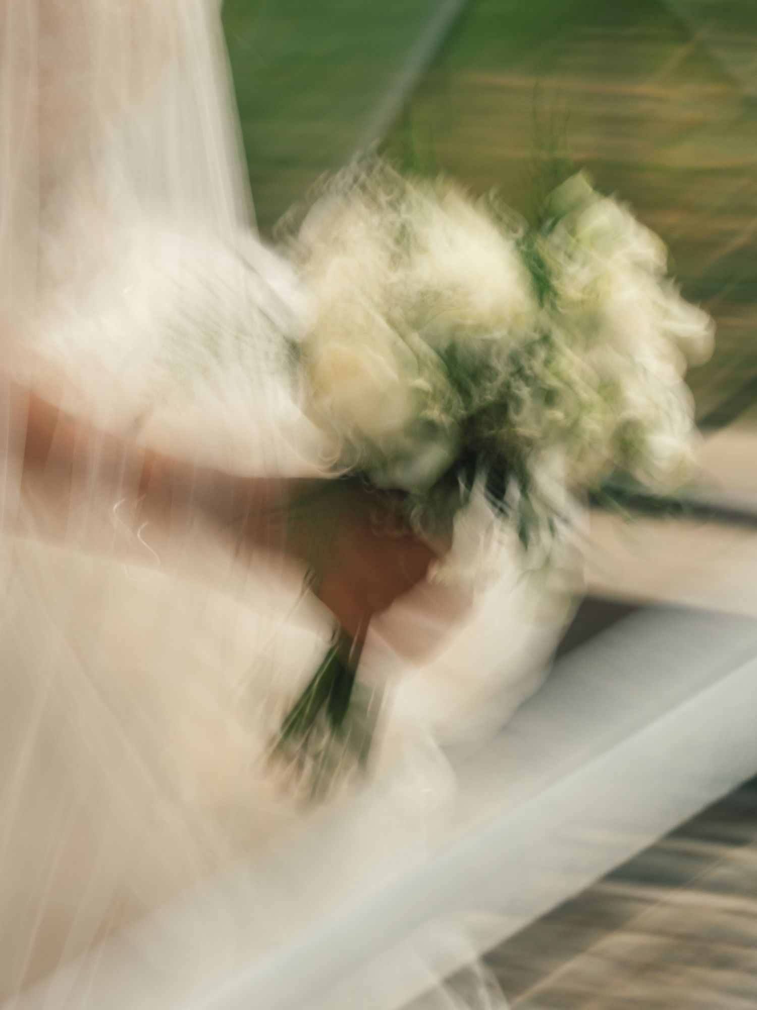 Blurry artistic shot of a bride holding a white and green wedding bouquet with veil flowing in foreground.