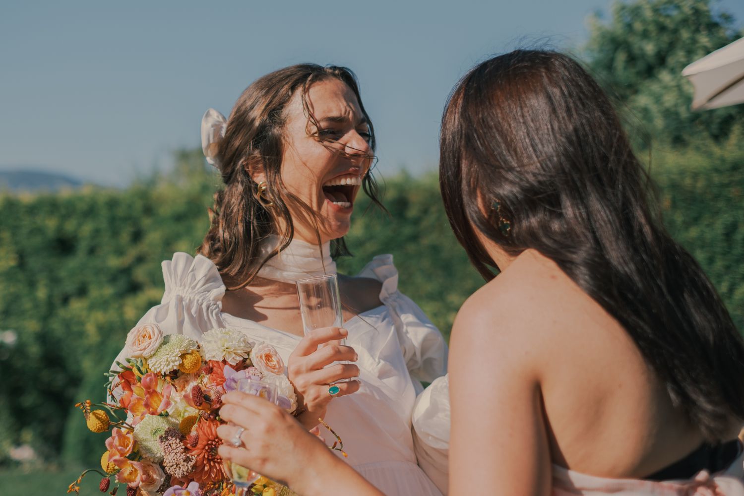 Joyful bride laughing with bridesmaid while holding pink drink and colorful bouquet outdoors.