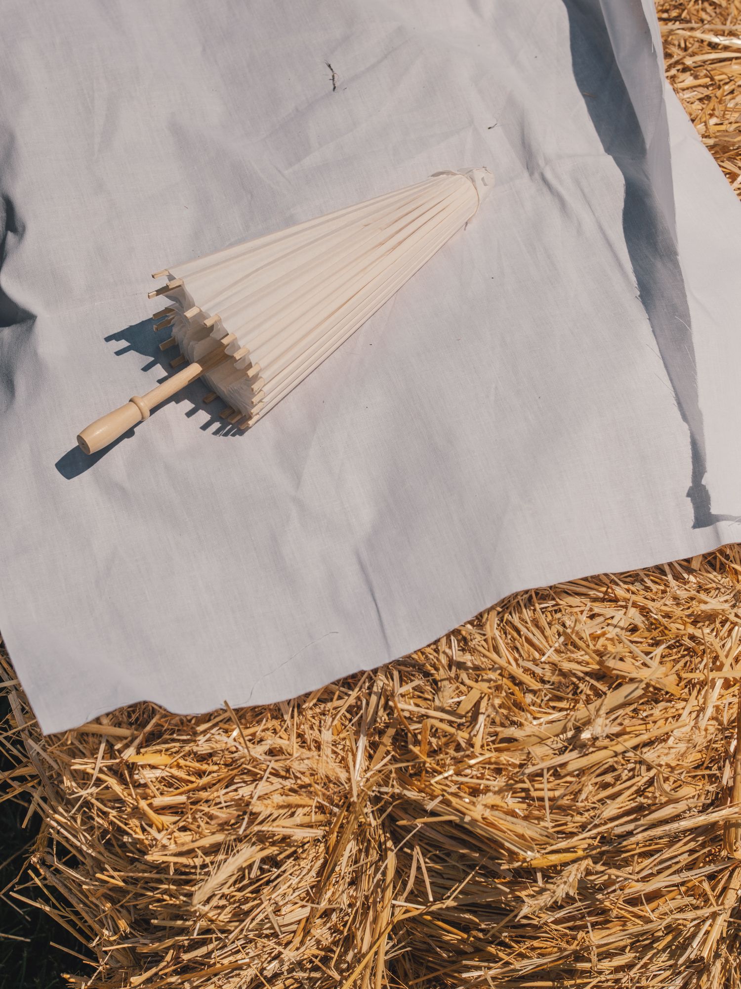 A cream parasol with wooden handle resting on a gray cloth draped over a rustic hay bale outdoors.