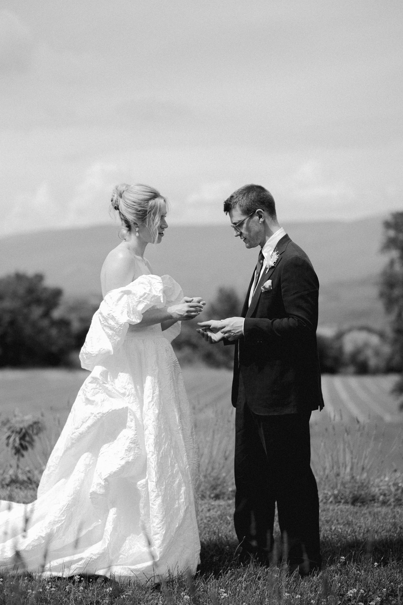 Black and white photo of a bride and groom exchanging rings outdoors with scenic mountain views behind them.