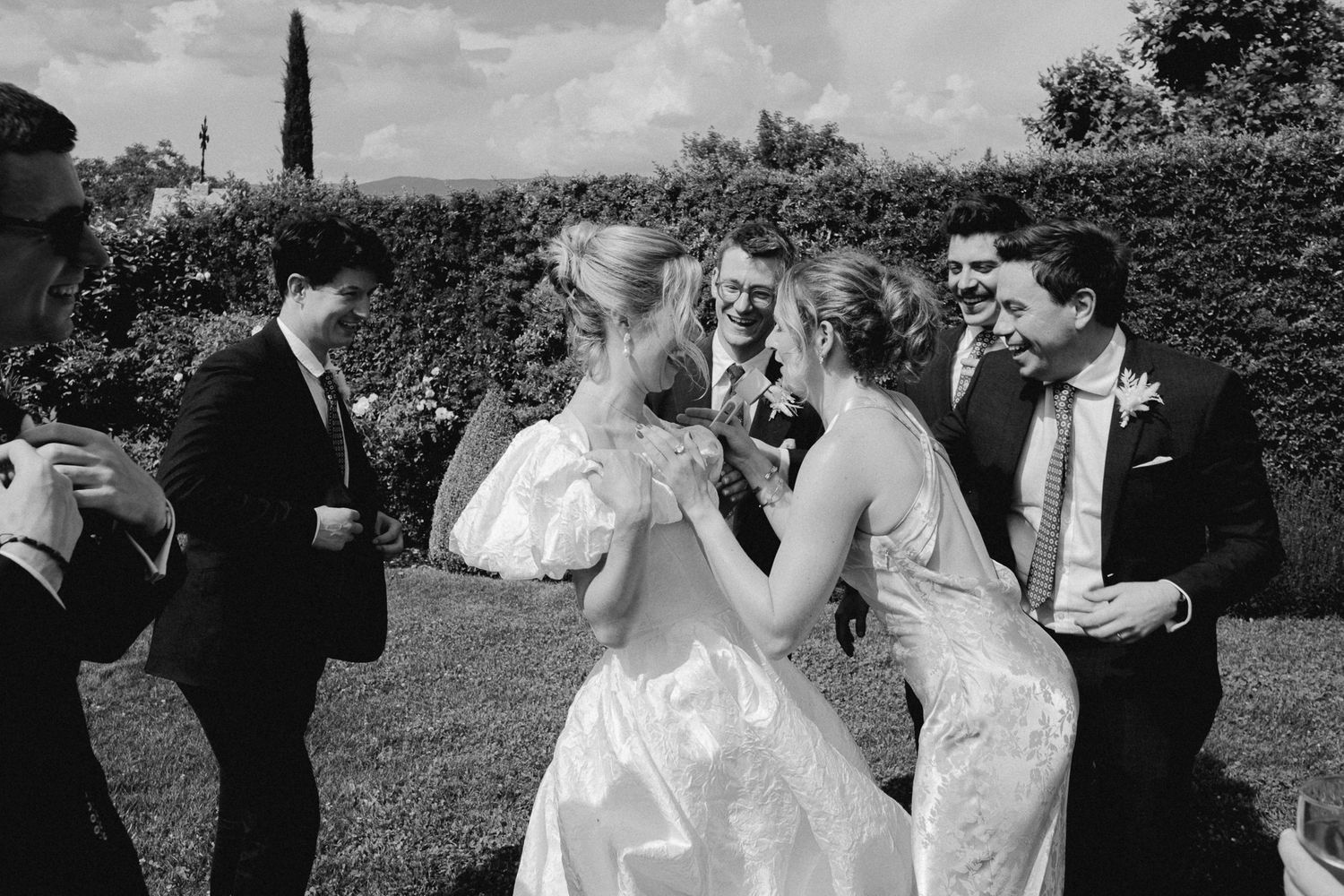Black and white photo of a bride and bridesmaids laughing with groomsmen at an outdoor wedding reception.