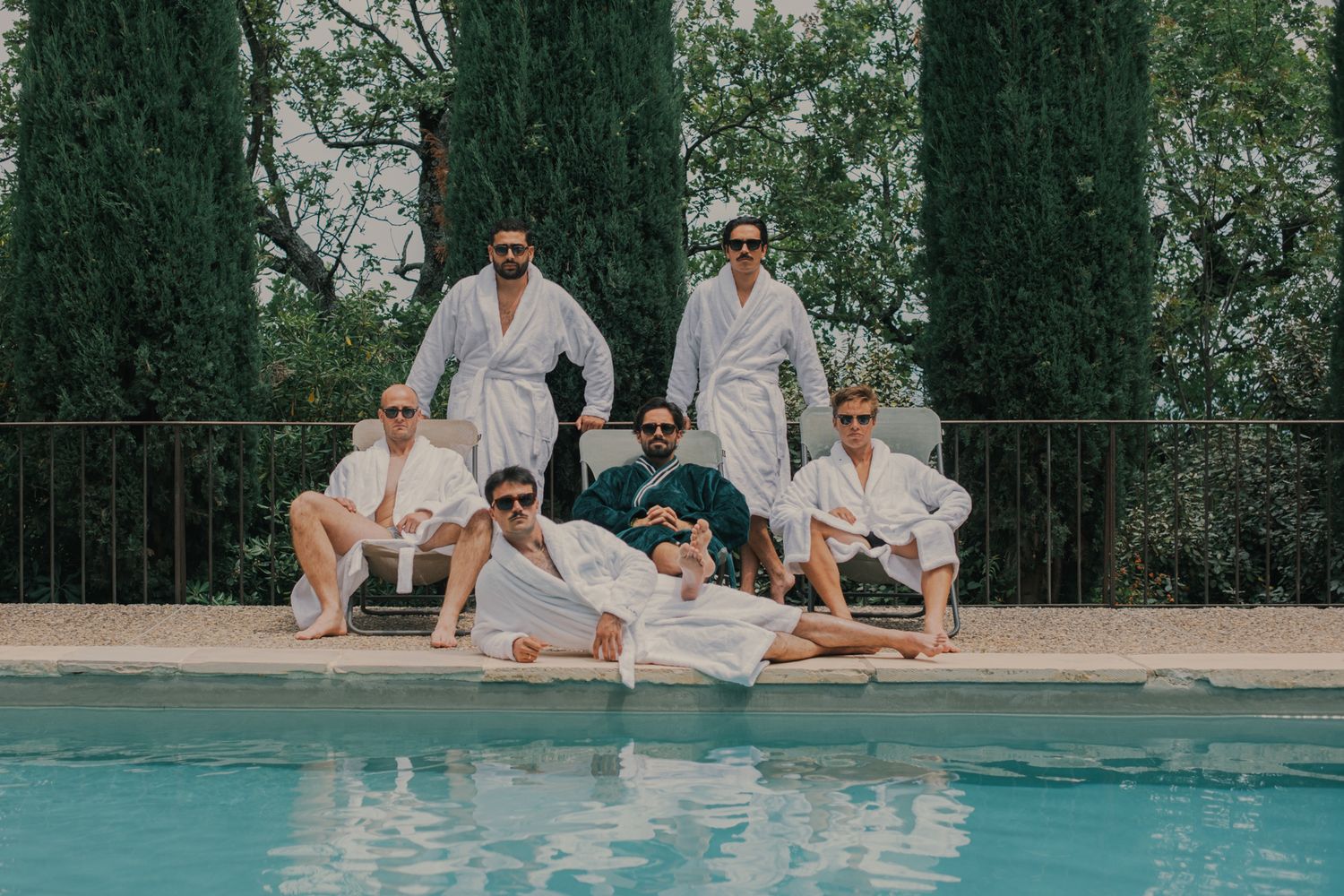 Six men in white robes posing poolside with cypress trees in background at a luxury resort.