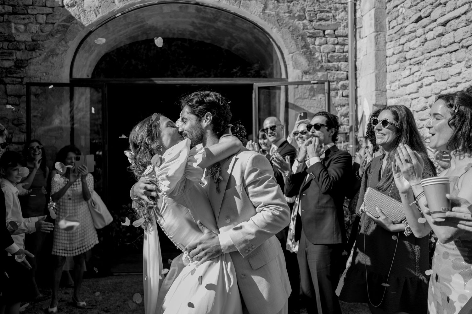Black and white photo of a couple sharing a kiss outside a stone building during their wedding celebration.