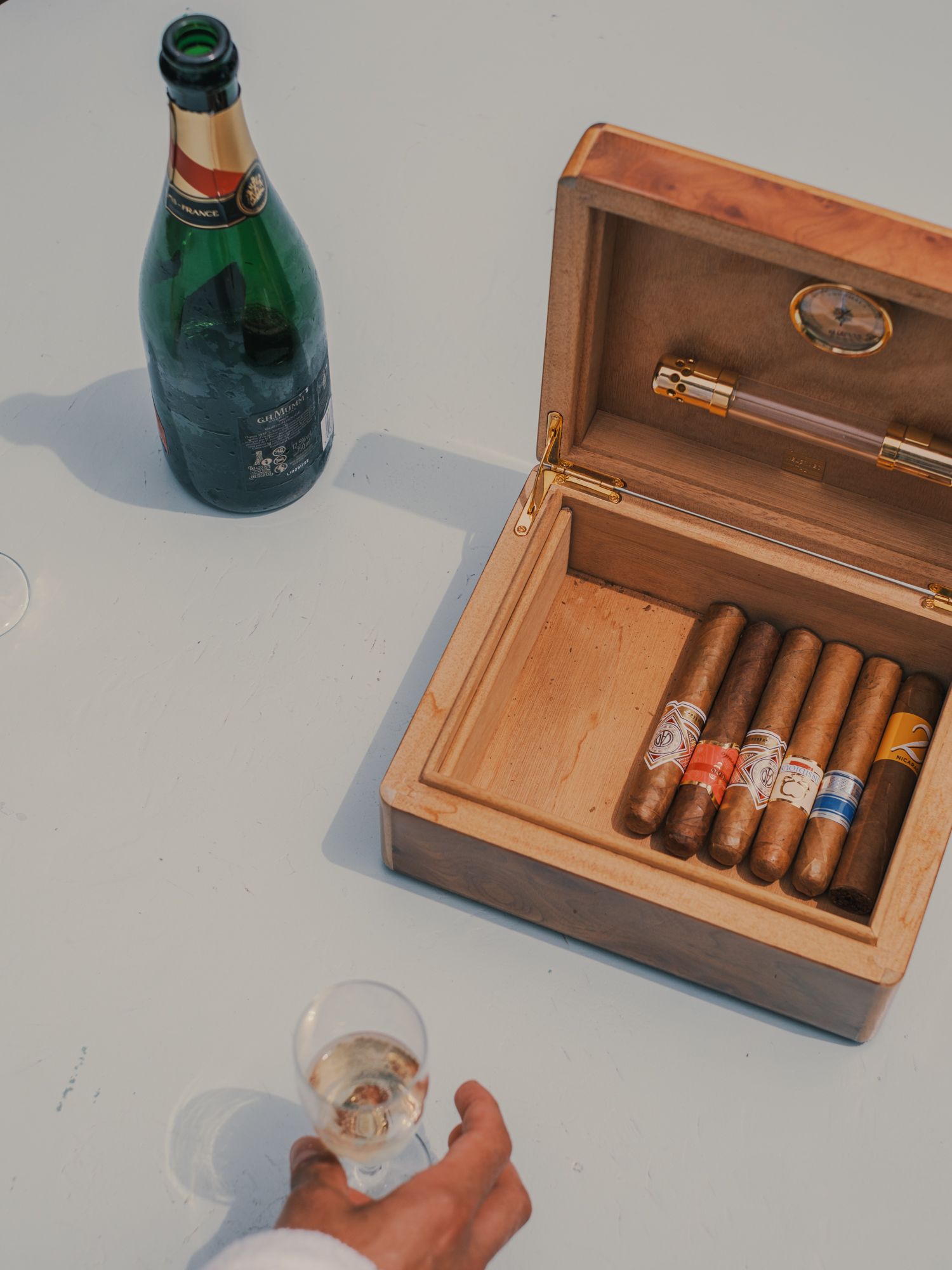 Open wooden humidor with assorted cigars beside a champagne bottle and glass on a white table.