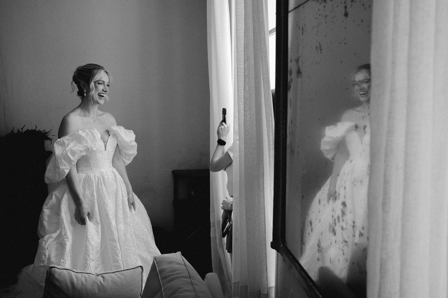 Black and white photo of a smiling bride in a puff-sleeve wedding gown reflected in a mirror.
