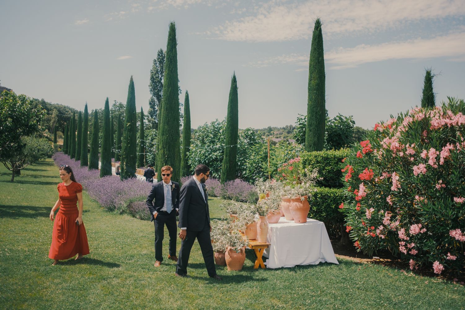 Elegant outdoor wedding reception in a Tuscan garden with cypress trees, lavender, and guests mingling near a decorated table.