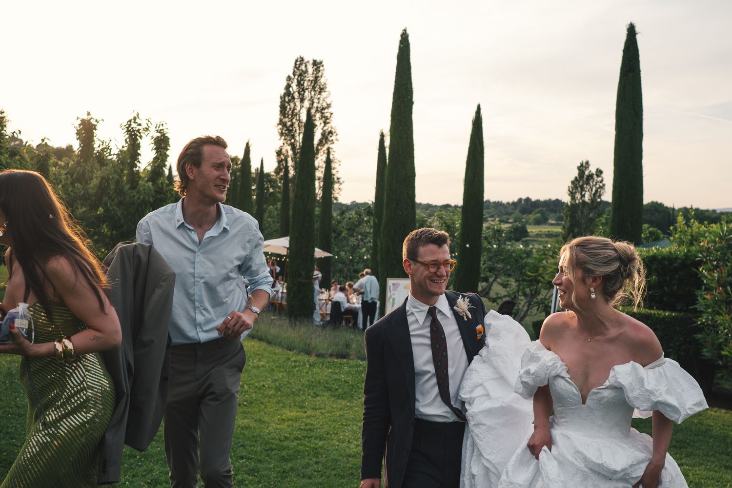 Bride in white off-shoulder gown laughs with groom and guests at outdoor wedding reception with cypress trees.