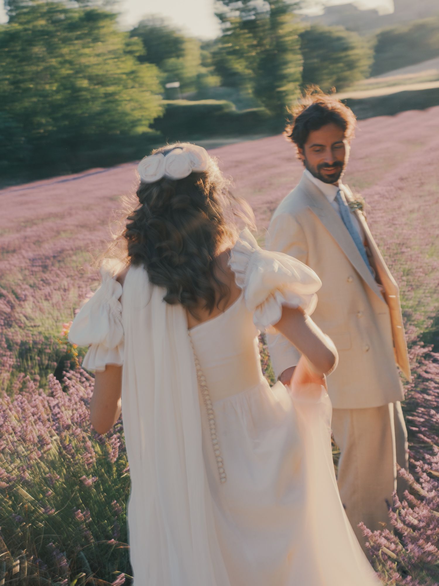 A bride and groom walk hand in hand through a blooming lavender field, she in a white gown, he in a beige suit.