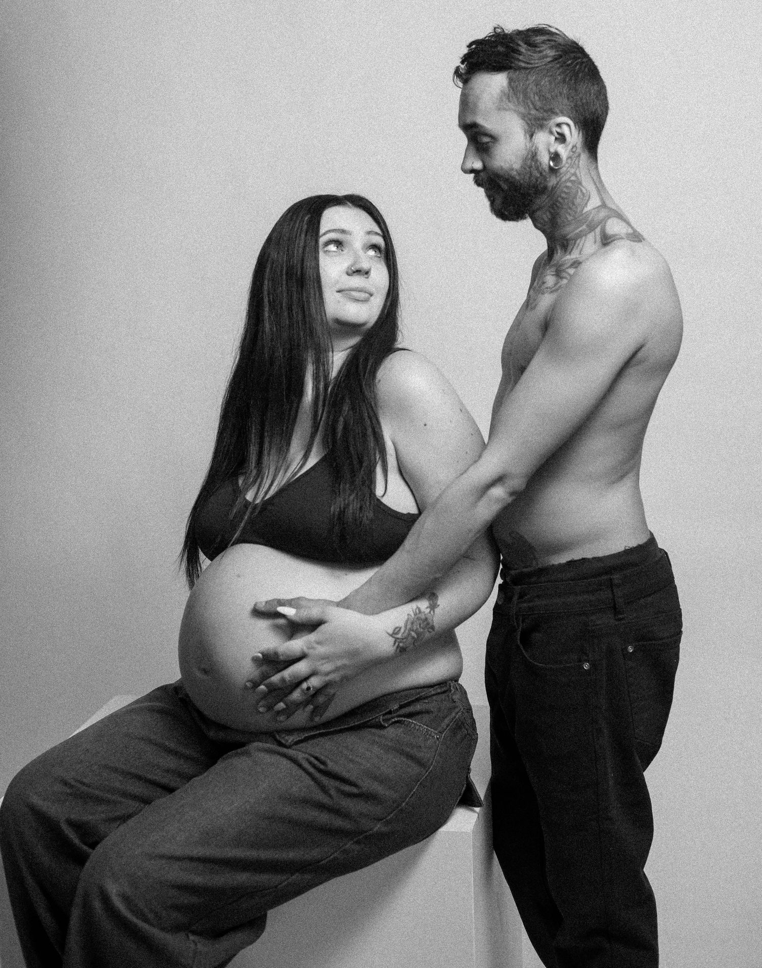Black and white maternity photo of pregnant woman seated with shirtless tattooed partner beside her.