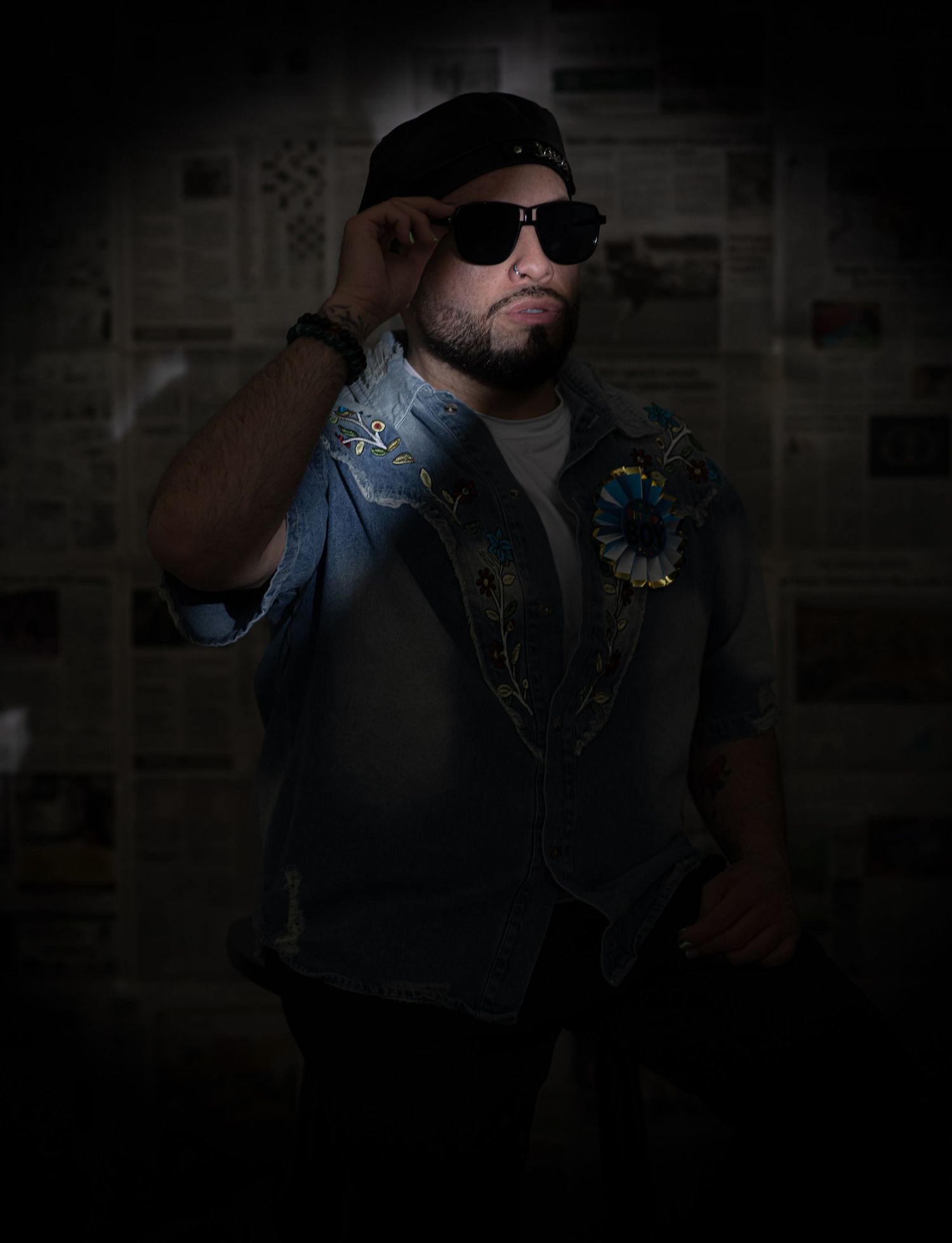 Man in sunglasses and embroidered denim jacket adjusting cap against newspaper backdrop.