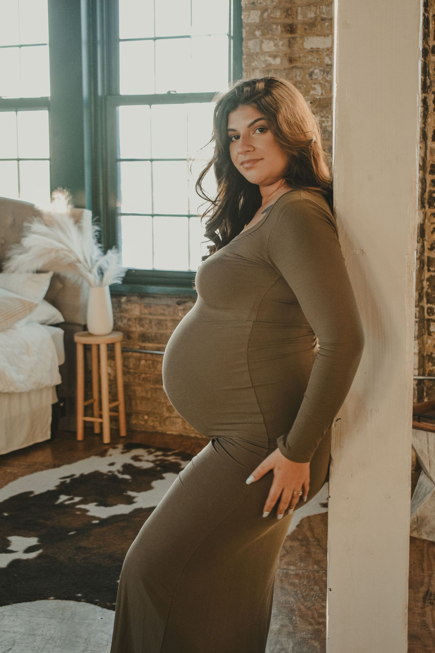 Pregnant woman in olive green form-fitting dress leaning against white pillar in rustic loft.