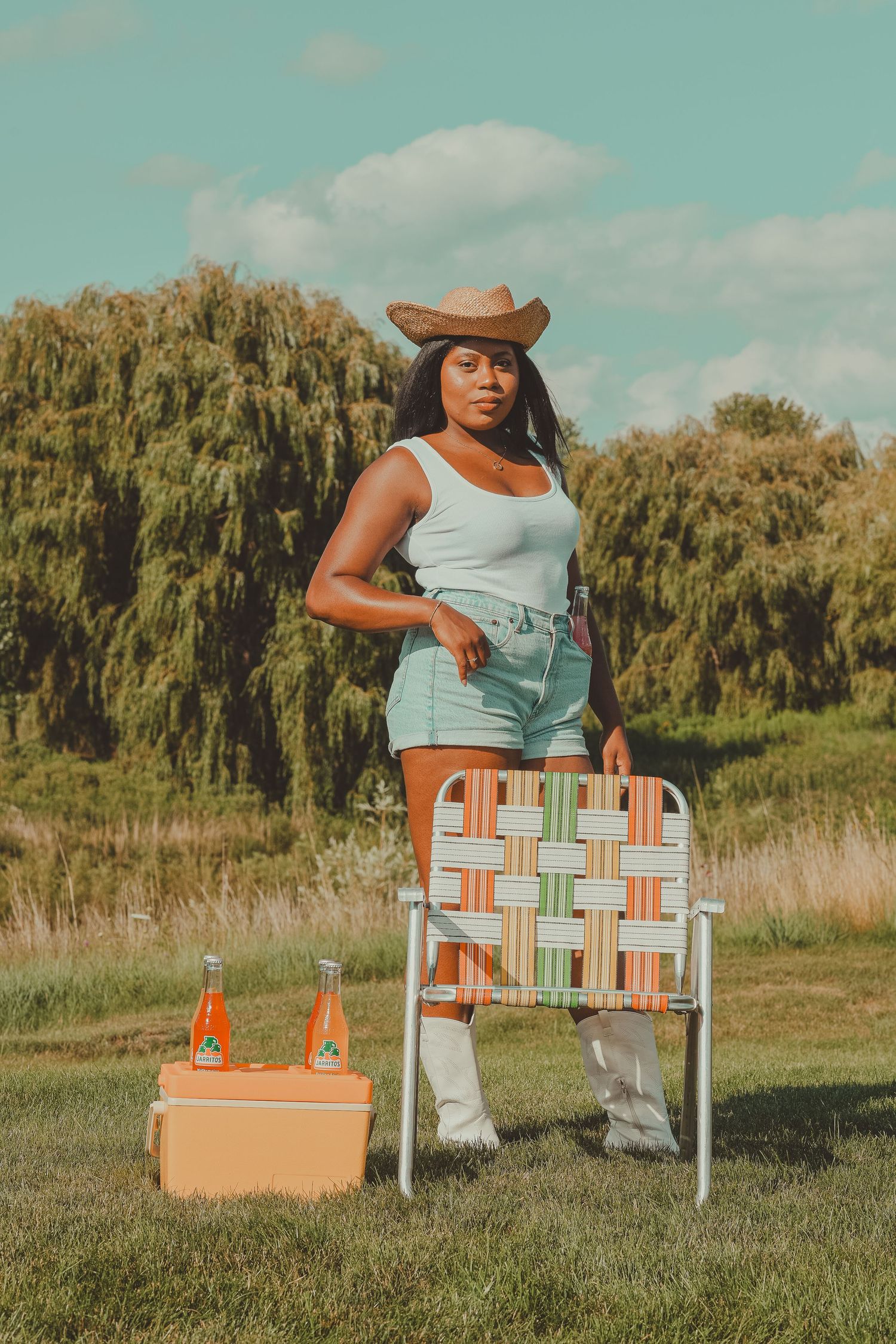 Woman in cowboy hat, white tank top, and denim shorts standing beside retro lawn chair outdoors.
