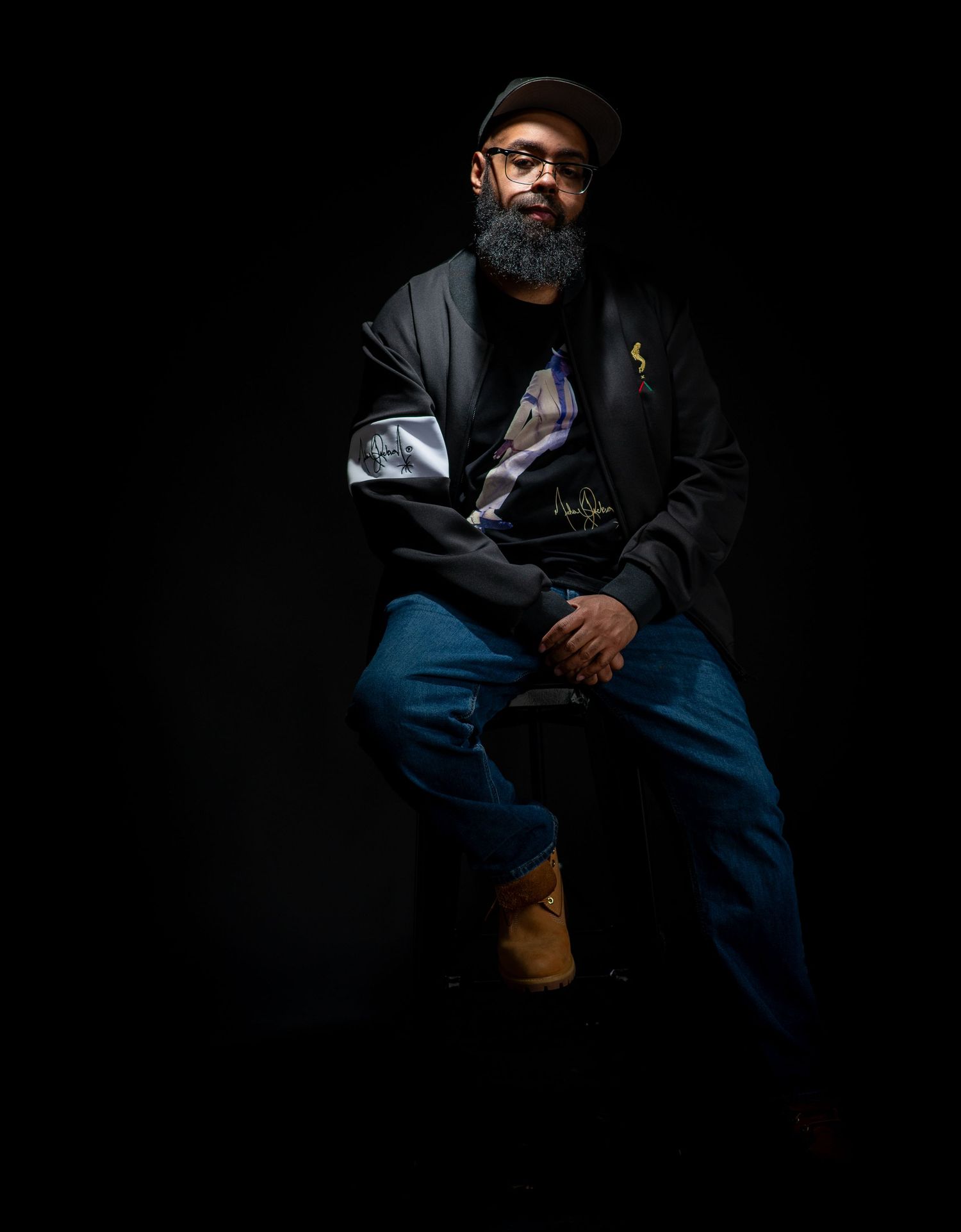 A bearded man in dark jacket and jeans sits confidently posed against a black background in dramatic lighting.