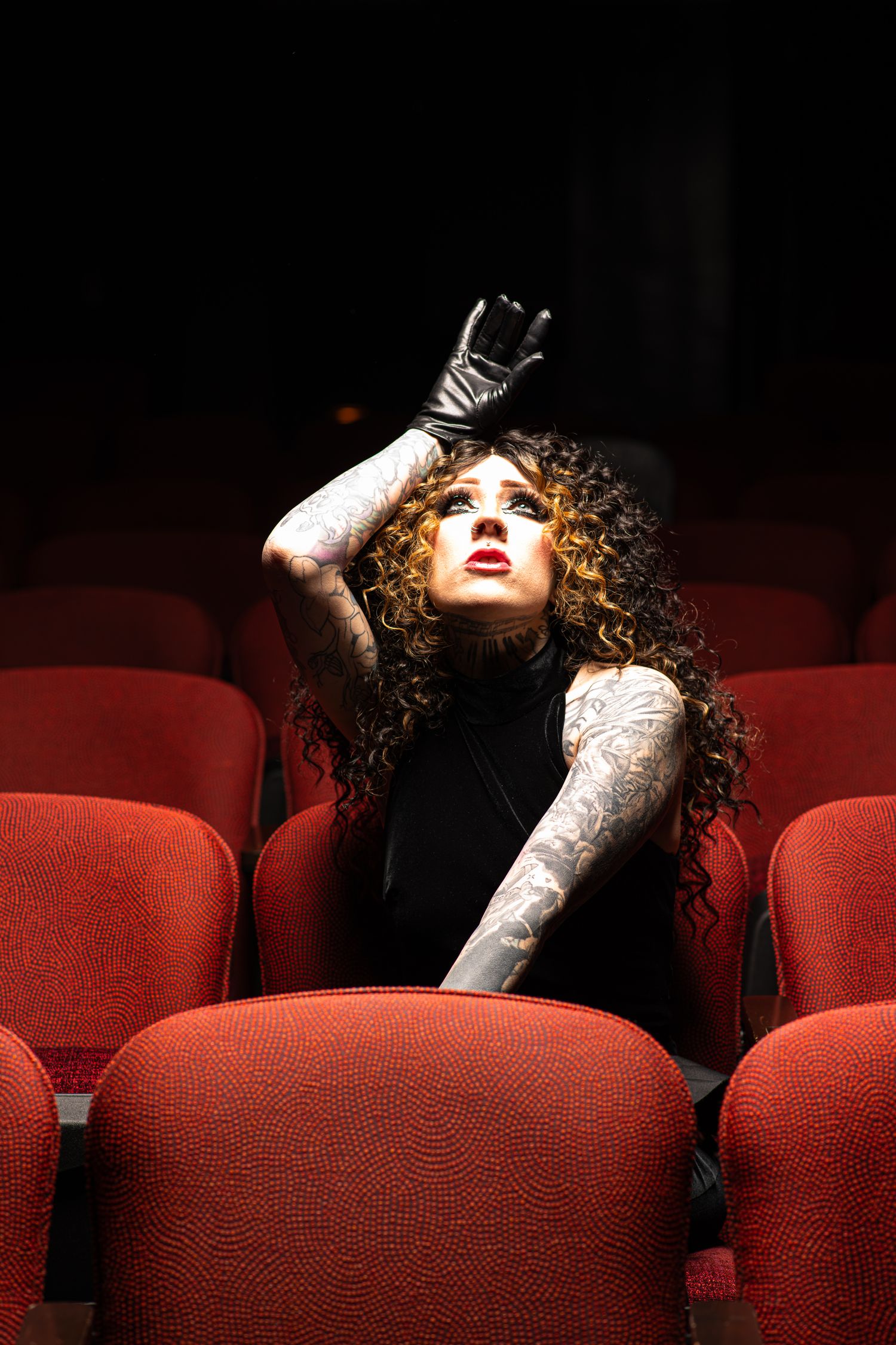 A tattooed woman with curly red hair in black outfit poses dramatically among red theater seats.
