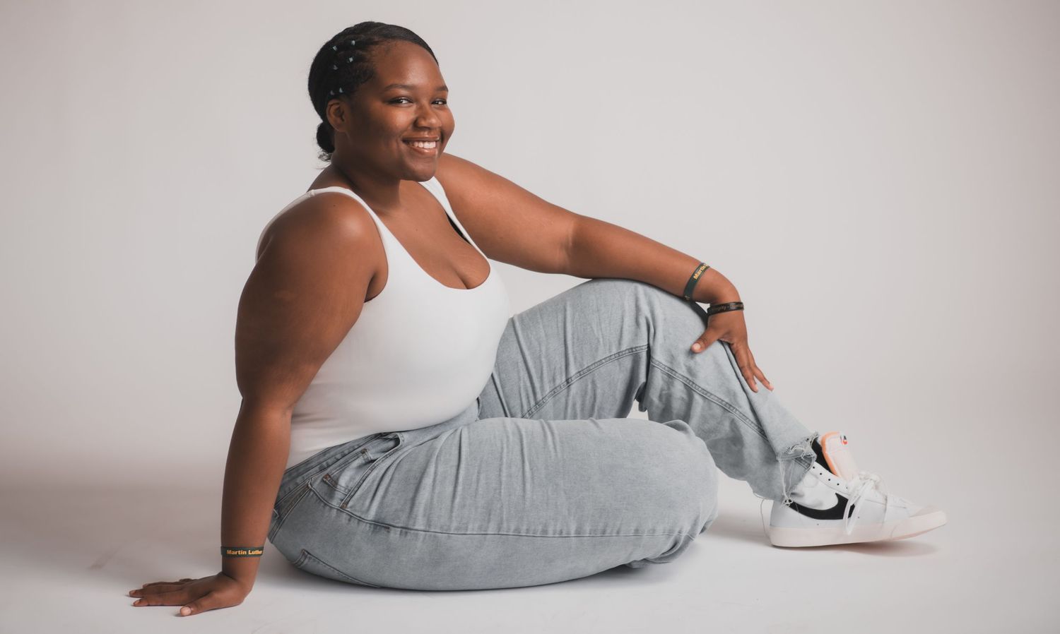 Female in white tank and jeans sitting casually on floor against gray background.