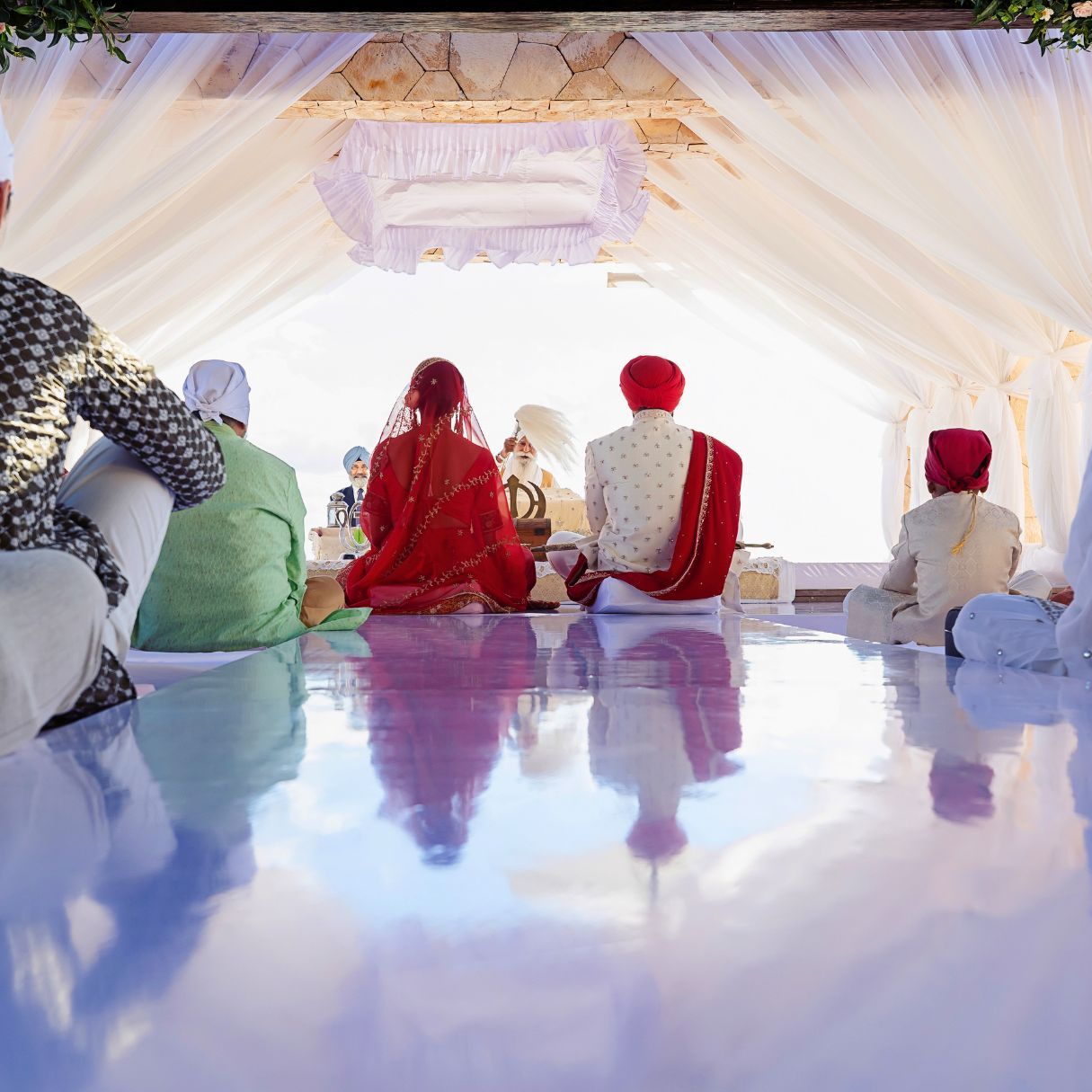 Sikh wedding ceremony with bride and groom seated under decorated mandap, viewed from behind with reflective floor.