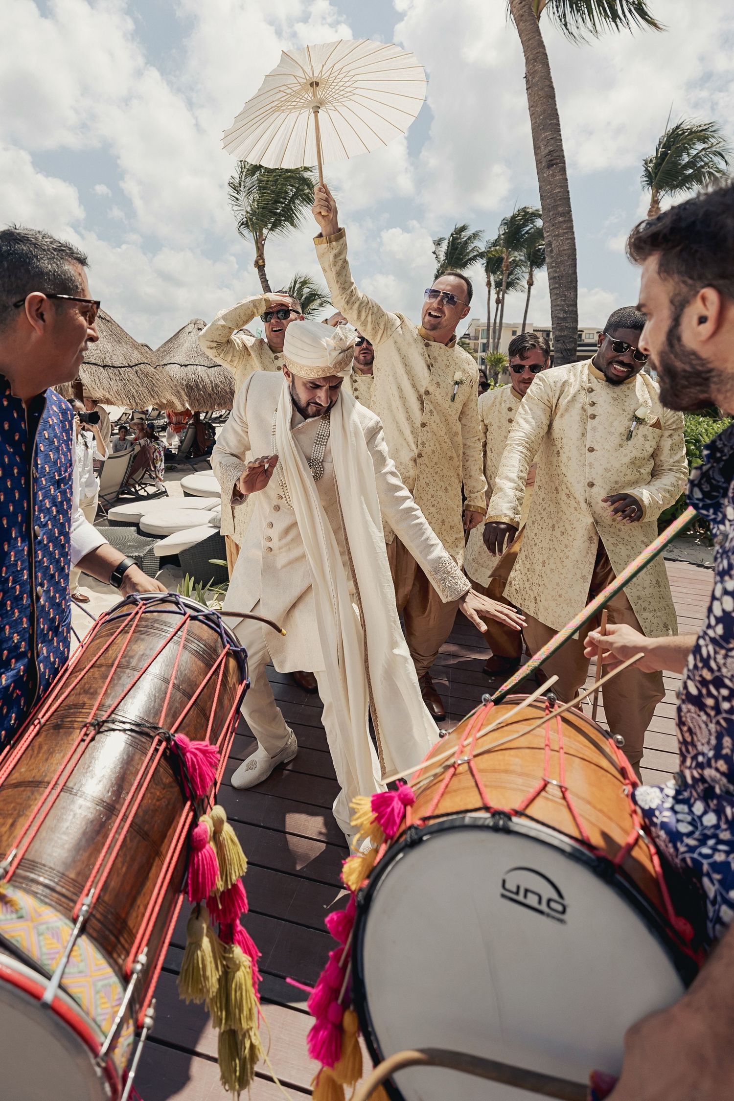 Groom in white sherwani dances with umbrella during Indian wedding baraat ceremony with dhol drummers outdoors.