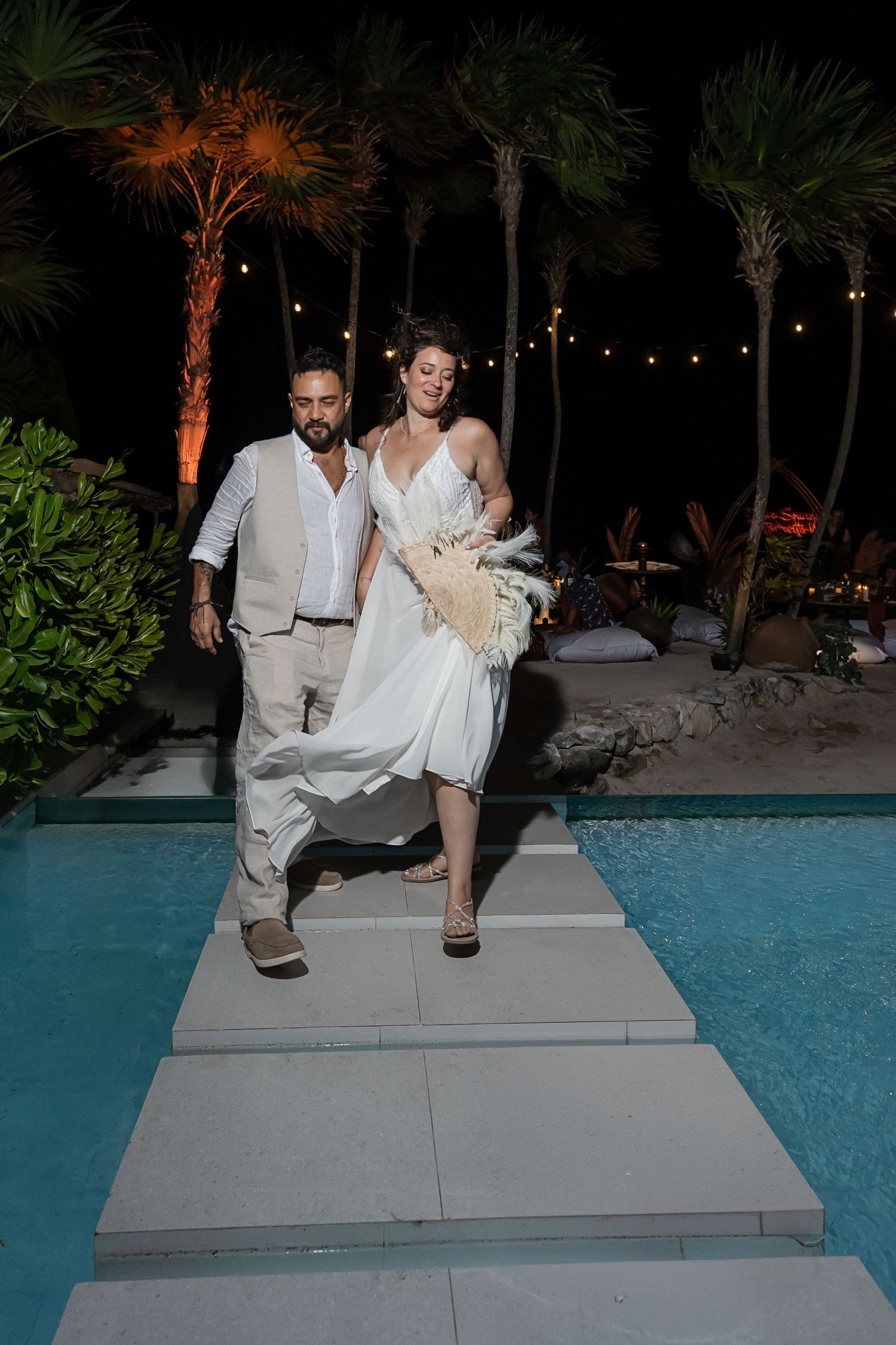 Bride and groom walking by a pool at night, bride in white dress holding bouquet, surrounded by palm trees and string lights.