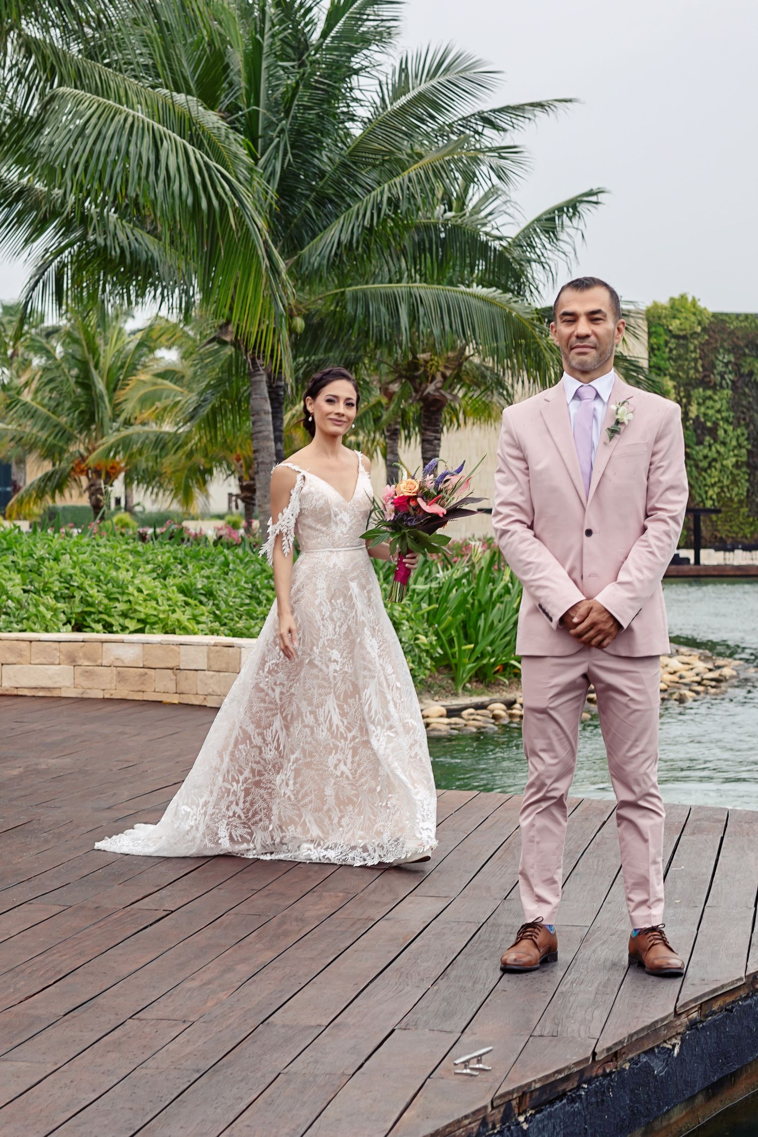 A couple in elegant wedding attire poses on a wooden deck with tropical palm trees and water in the background.