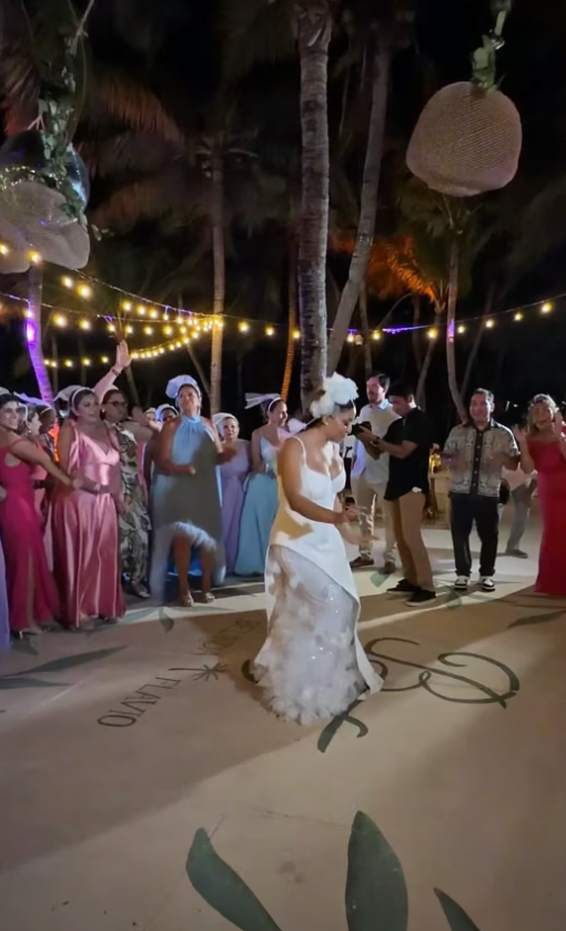 Bride in white gown tosses bouquet at outdoor evening wedding reception with string lights and palm trees.