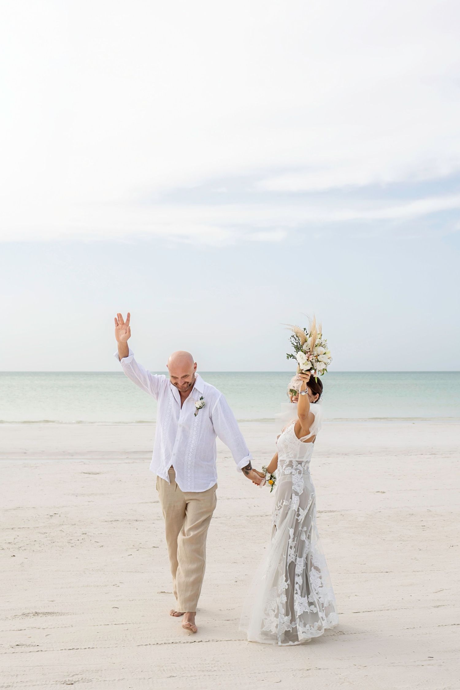 Newlywed couple in white attire celebrating on a sandy beach, holding hands with ocean backdrop.