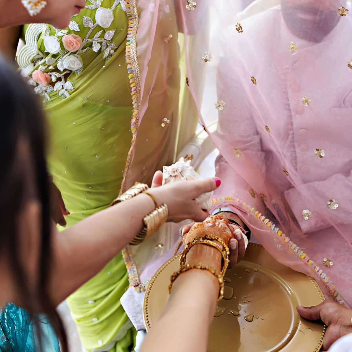 Women in colorful Indian attire perform a traditional wedding ritual, adorning a bride's hands with gold bangles and henna.