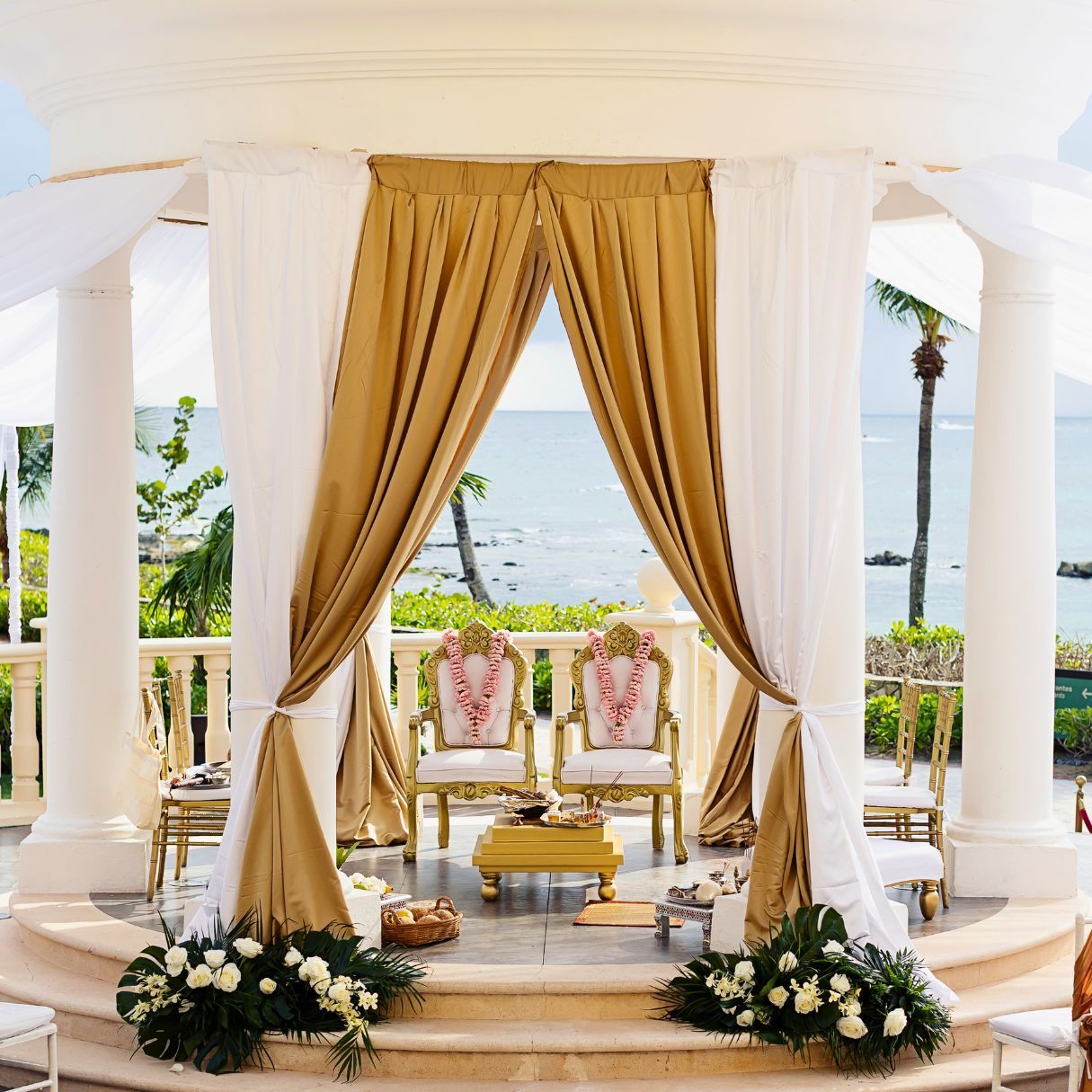 Elegant outdoor wedding gazebo with gold drapes, white columns, ornate chairs, floral arrangements, and ocean view backdrop.