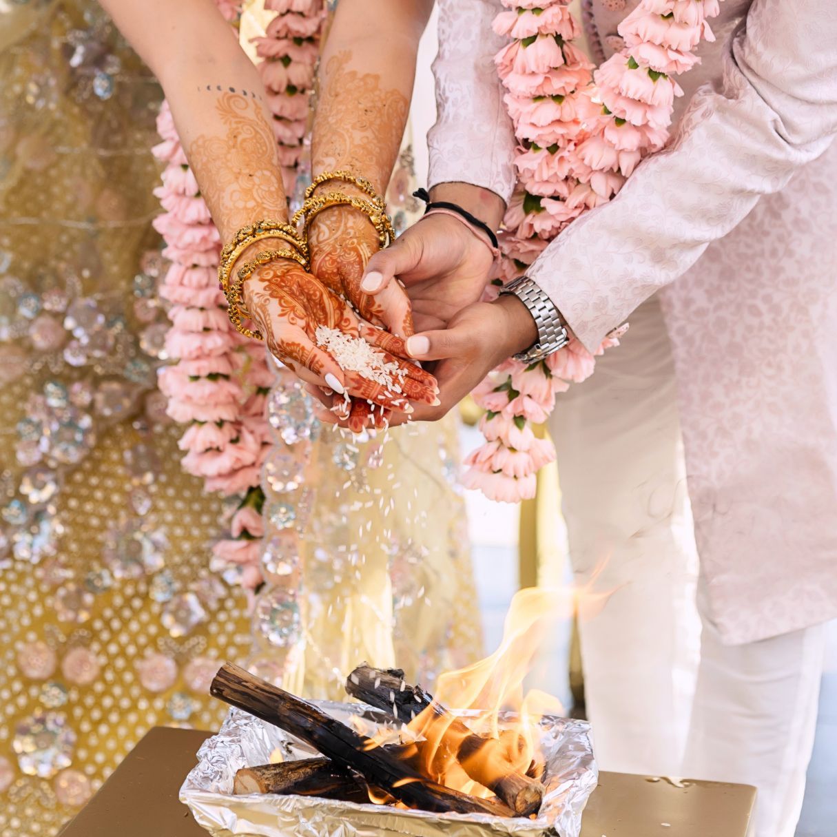 Bride and groom performing Hindu wedding fire ritual, pouring offerings into sacred flame, adorned with floral garlands.