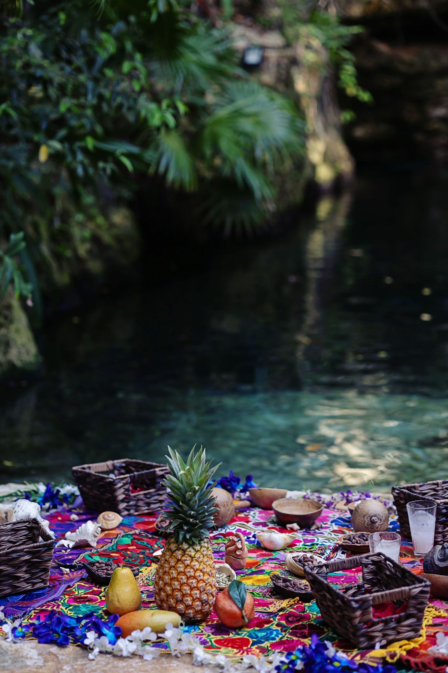 Colorful textiles and decorative objects including a pineapple displayed along the edge of a dark reflective pool or water.