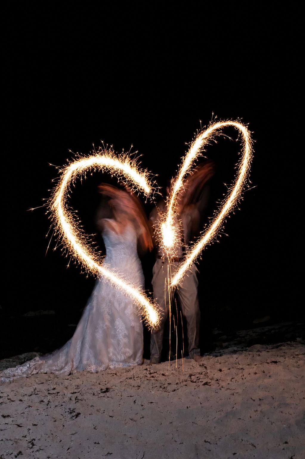 Couple in wedding attire creating a glowing sparkler heart shape on a dark beach at night.