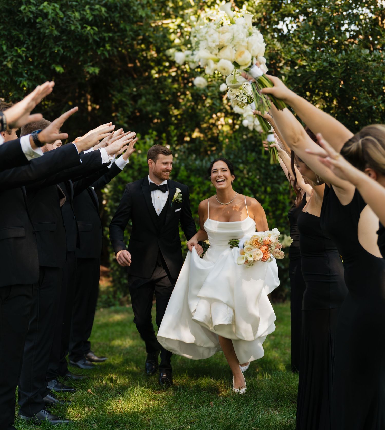 Wedding party walks together through an orchard at sunset with arms raised in celebration.