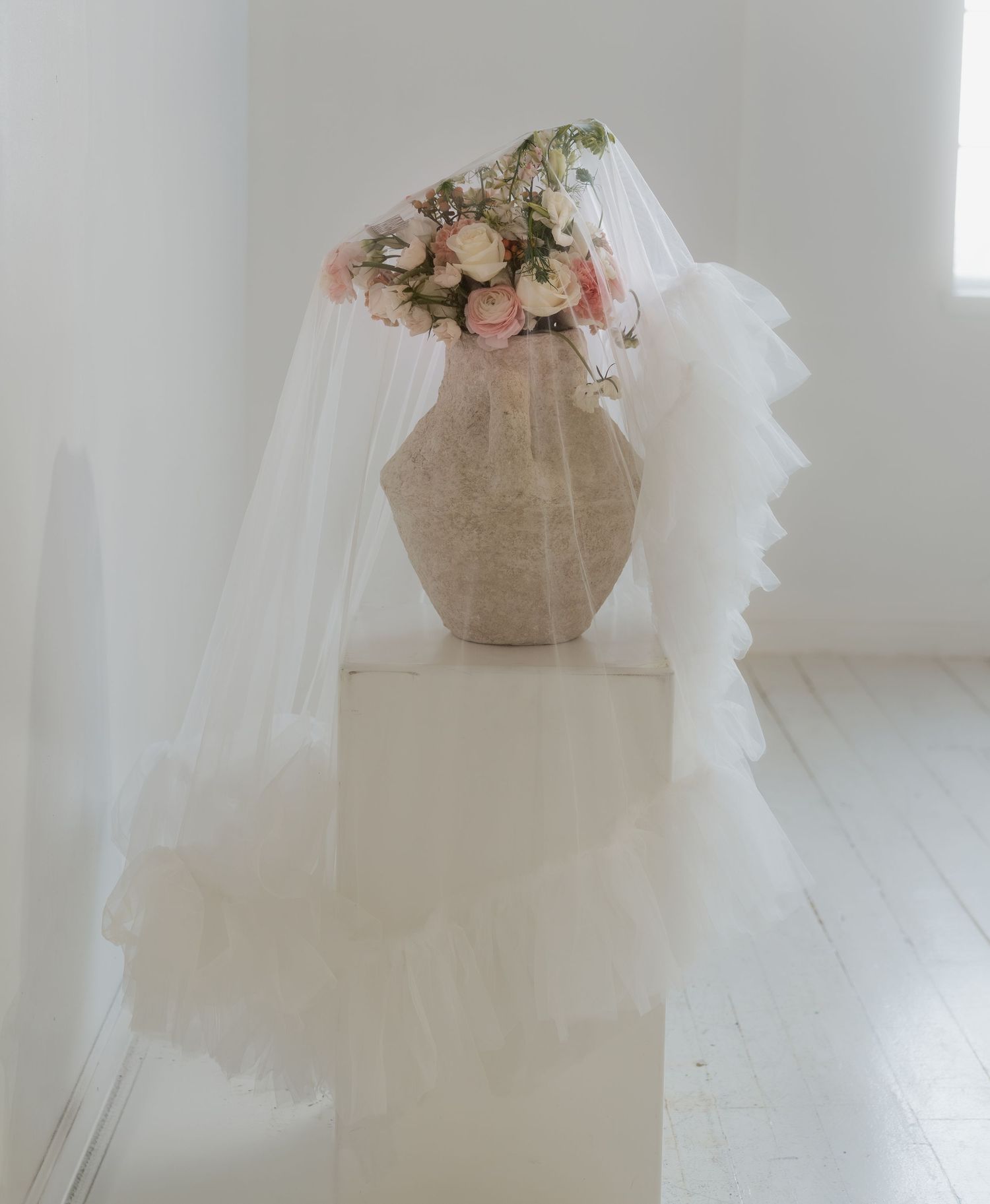 A delicate bridal veil and pink roses arranged on a stone pedestal against a white minimalist backdrop.