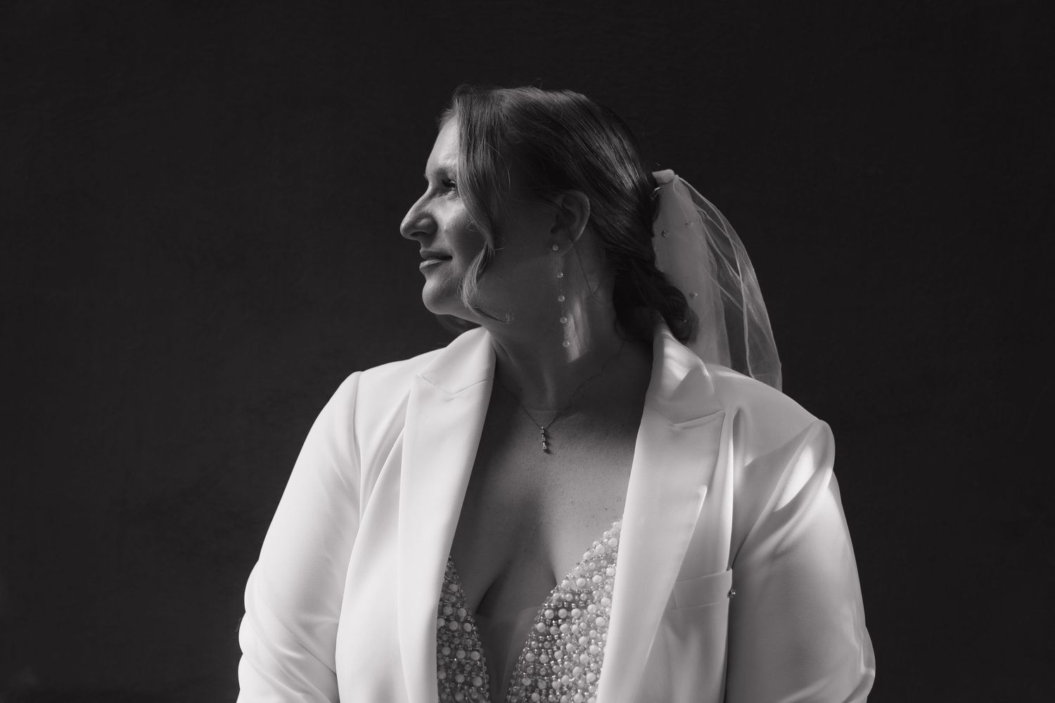 Black and white portrait of a smiling bride wearing a white blazer and embellished dress.