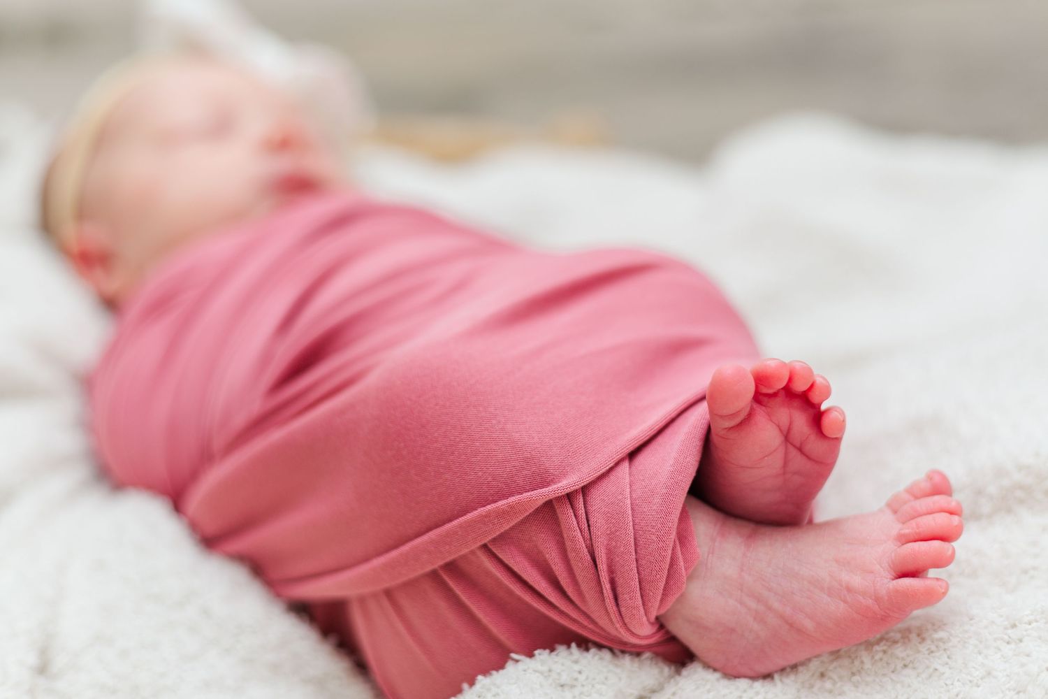 Baby wrapped in soft coral fabric lying on white blanket with tiny feet peeking out.