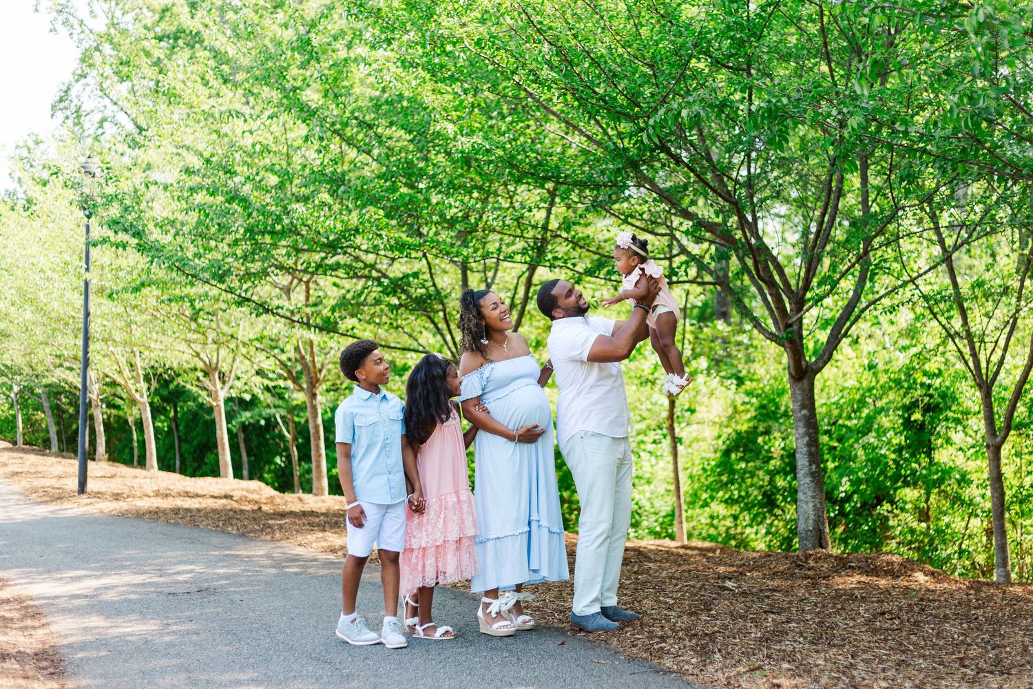 A joyful family in white outfits poses together in a lush green park, with a pregnant mother glowing.