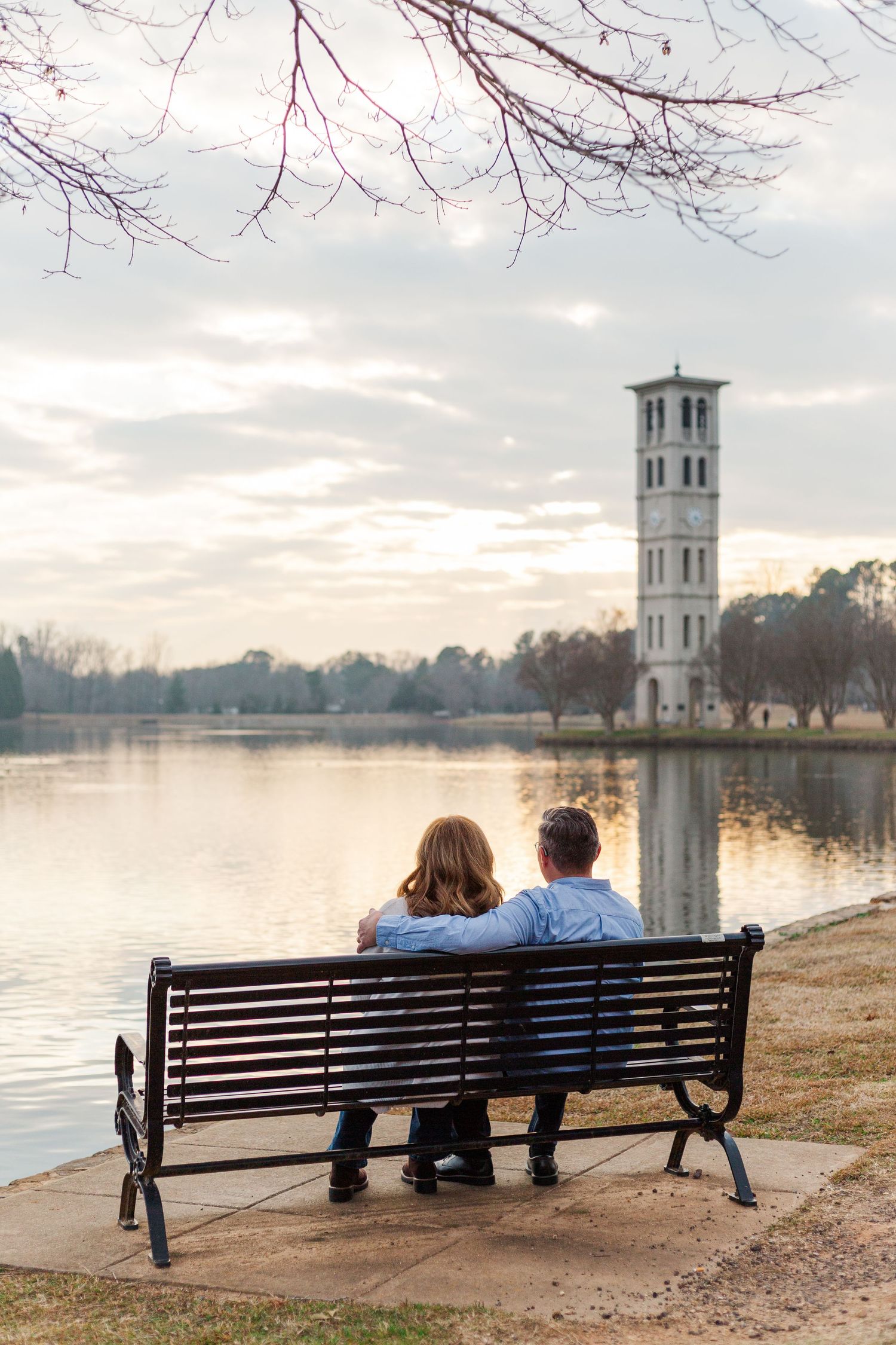 A couple sits on a bench overlooking a tranquil lake at sunset with a white bell tower in the background.