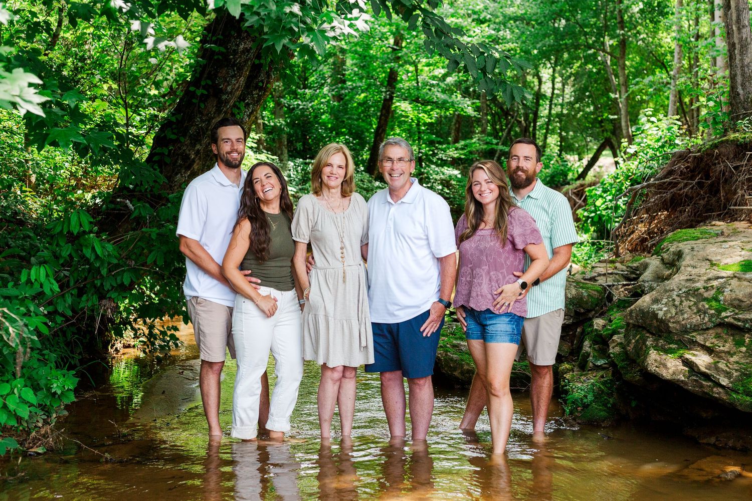 Extended family stands together in a shallow stream during an outdoor photo session in the woods.