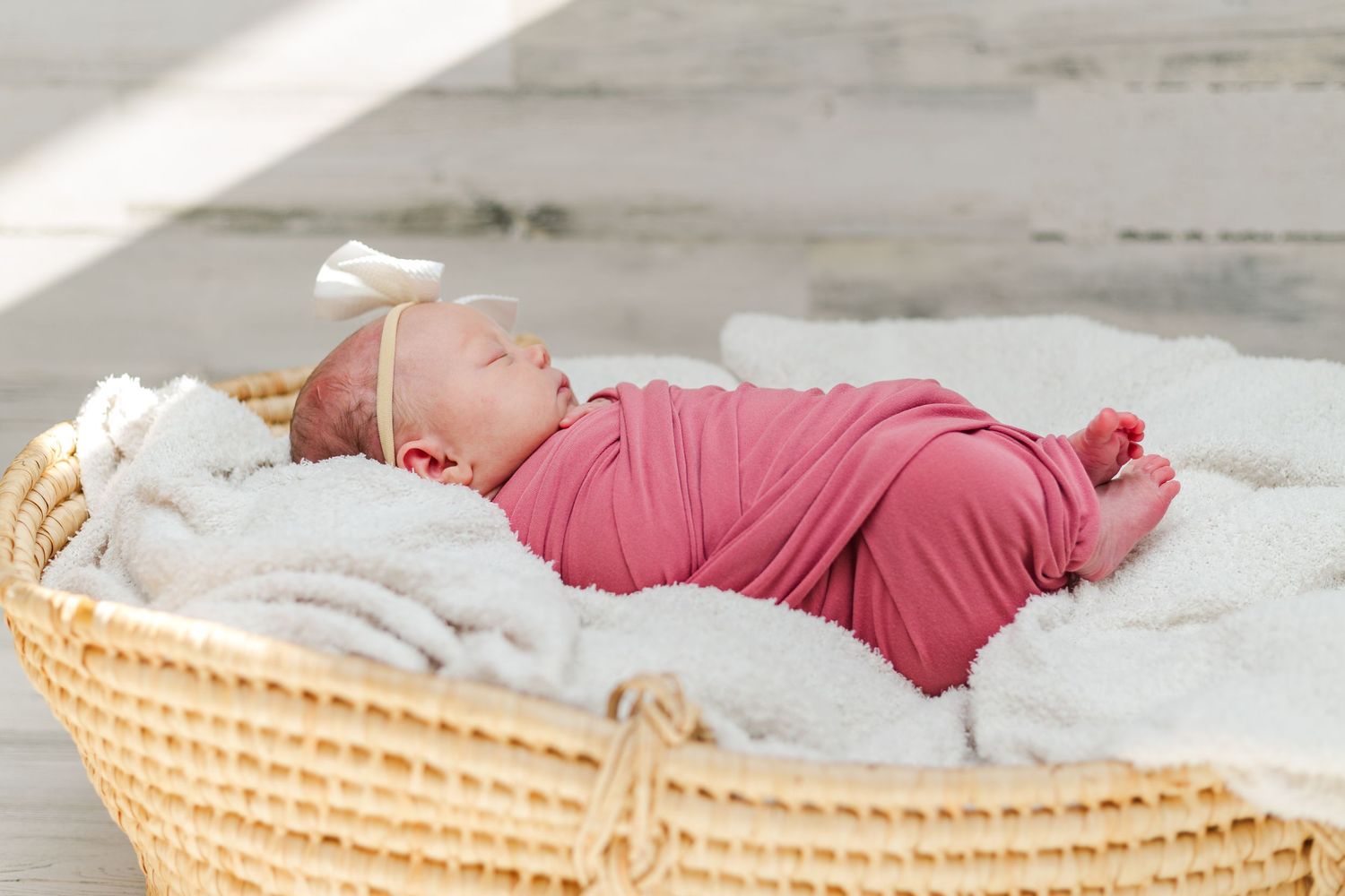 A sleeping newborn baby in burgundy clothing lies peacefully in a woven wicker basket on white bedding.