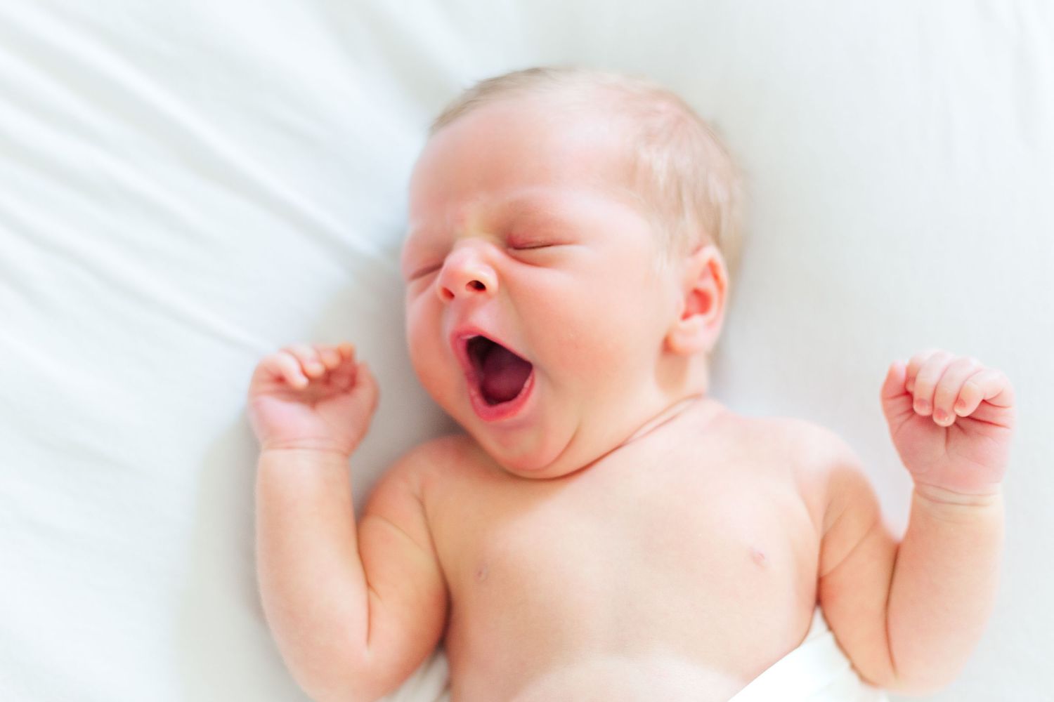 Newborn baby yawning and stretching arms while lying on white blanket wearing diaper.