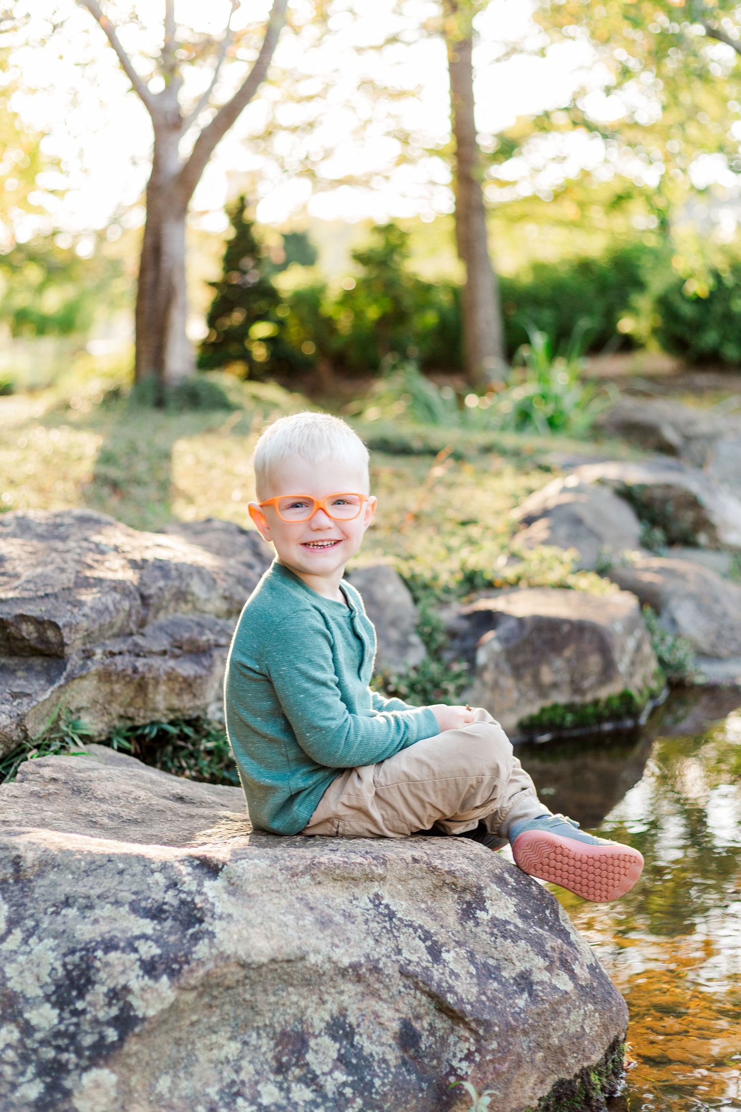 Child in green sweater and khaki pants sits on large rocks by a pond on a sunny autumn day.