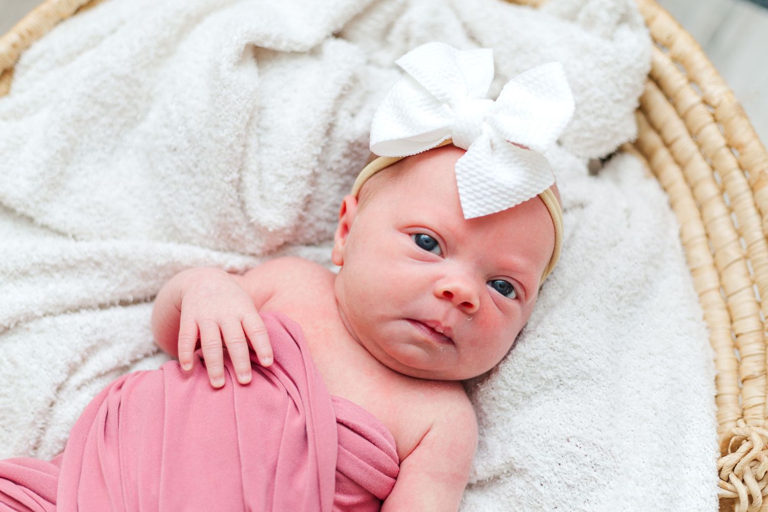 A newborn baby wearing a pink outfit and white bow headband lies on a cream-colored blanket.