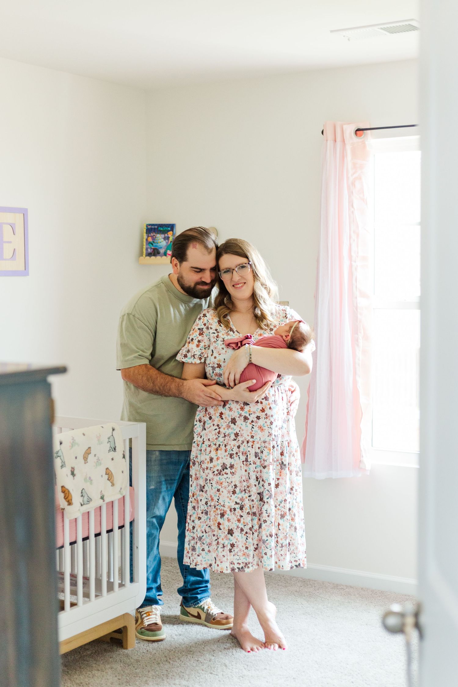 New parents proudly hold their newborn baby in a decorated nursery with white crib and pink accents.